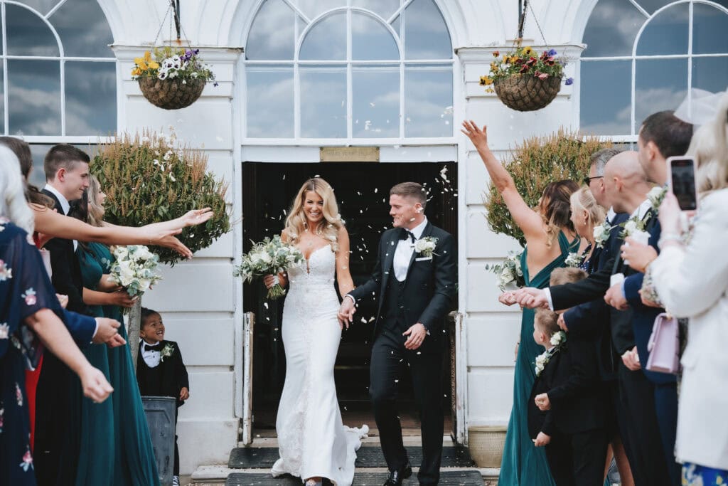 Bride and groom walking through a confetti shower as guests celebrate outside the Georgian entrance of Gosfield Hall, Essex — wedding photography by Lily & White