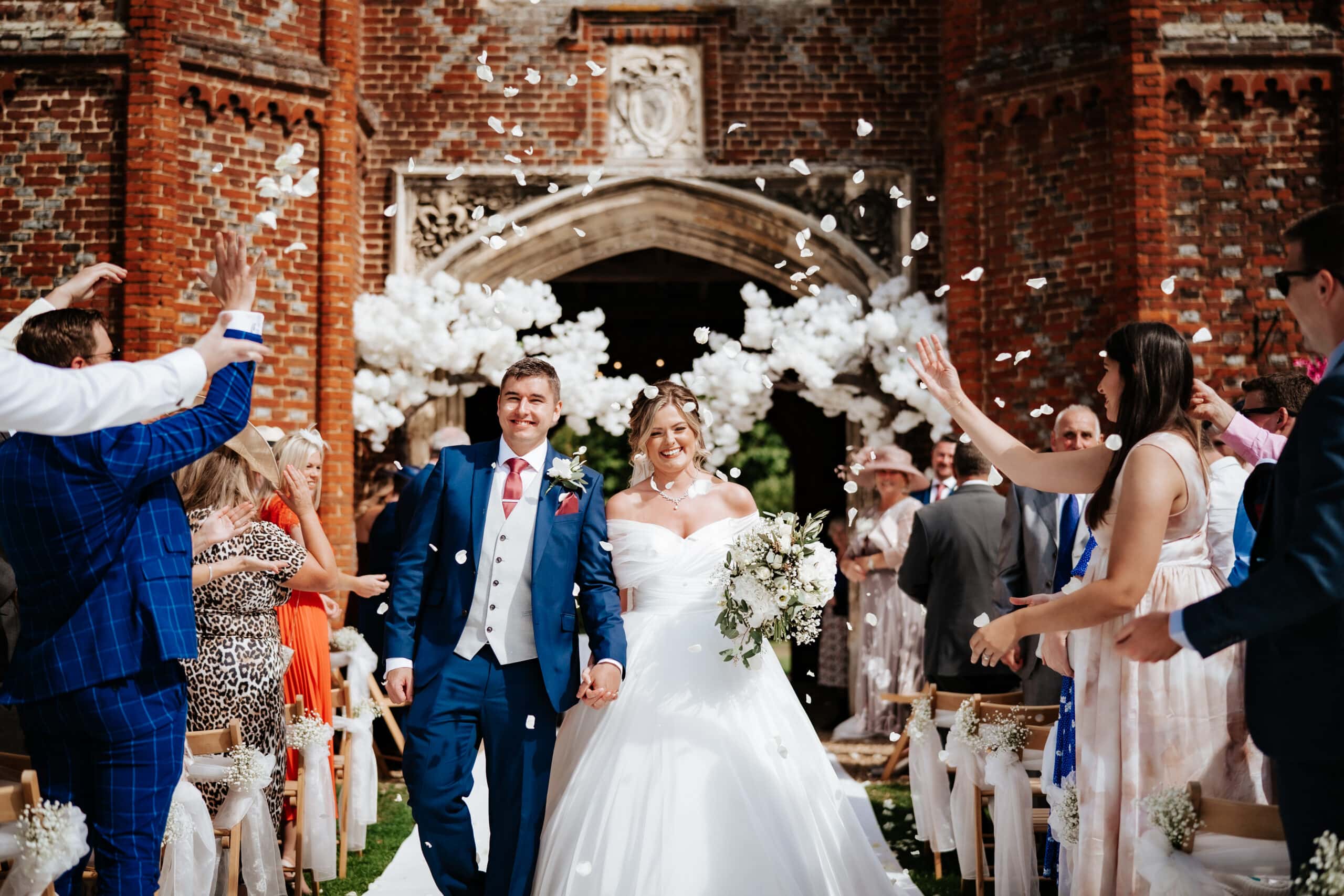 Bride and groom walking through confetti beneath the historic Tudor carriageway at Leez Priory wedding venue in Essex — joyful documentary wedding photography by Lily & White