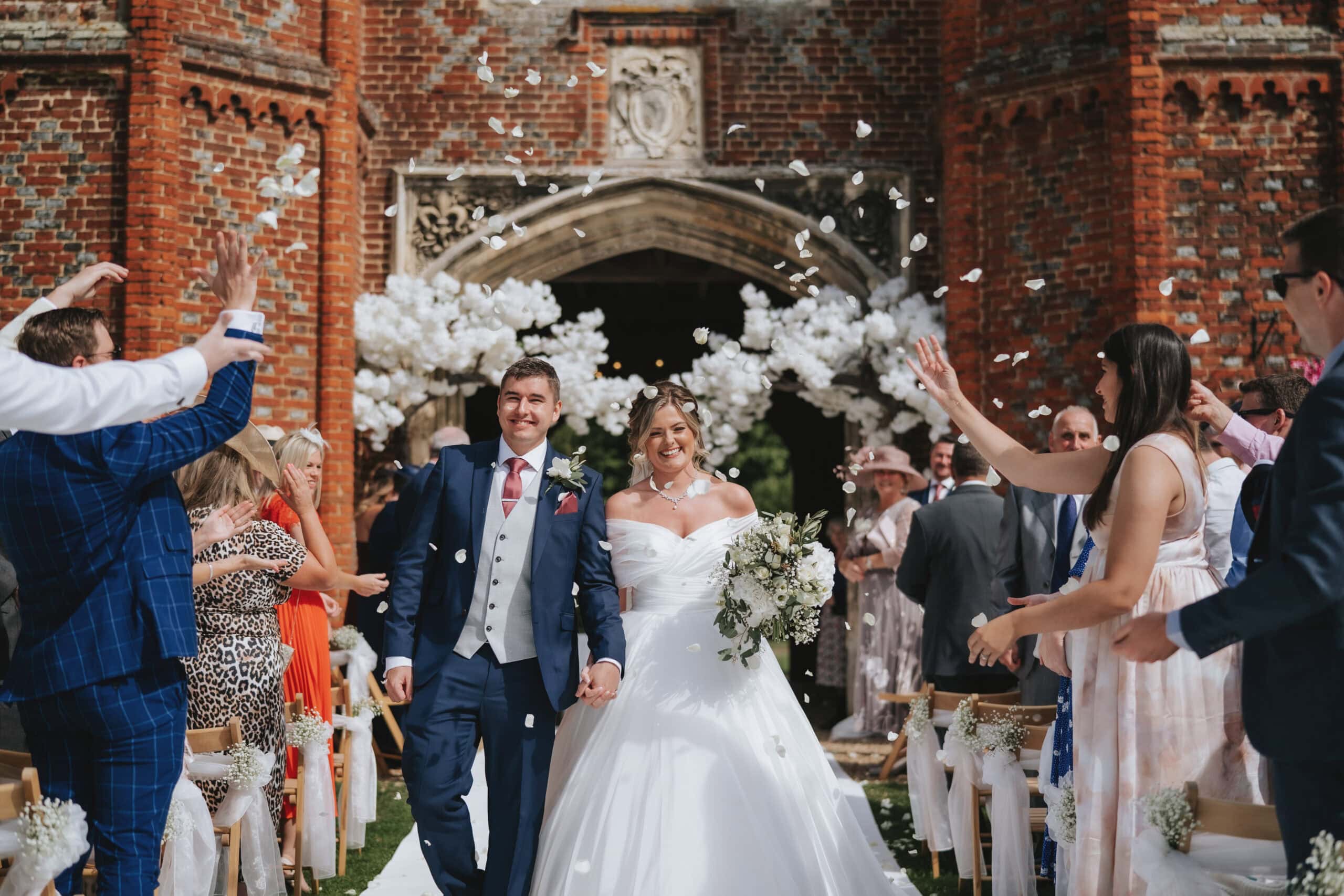 Rhiannon and Paul emerge through a flurry of white confetti beneath the historic Great Tower at Leez Priory. The ancient red brickwork of the Tudor tower creates a timeless, grand backdrop for this joyful, unscripted moment during their autumn wedding.