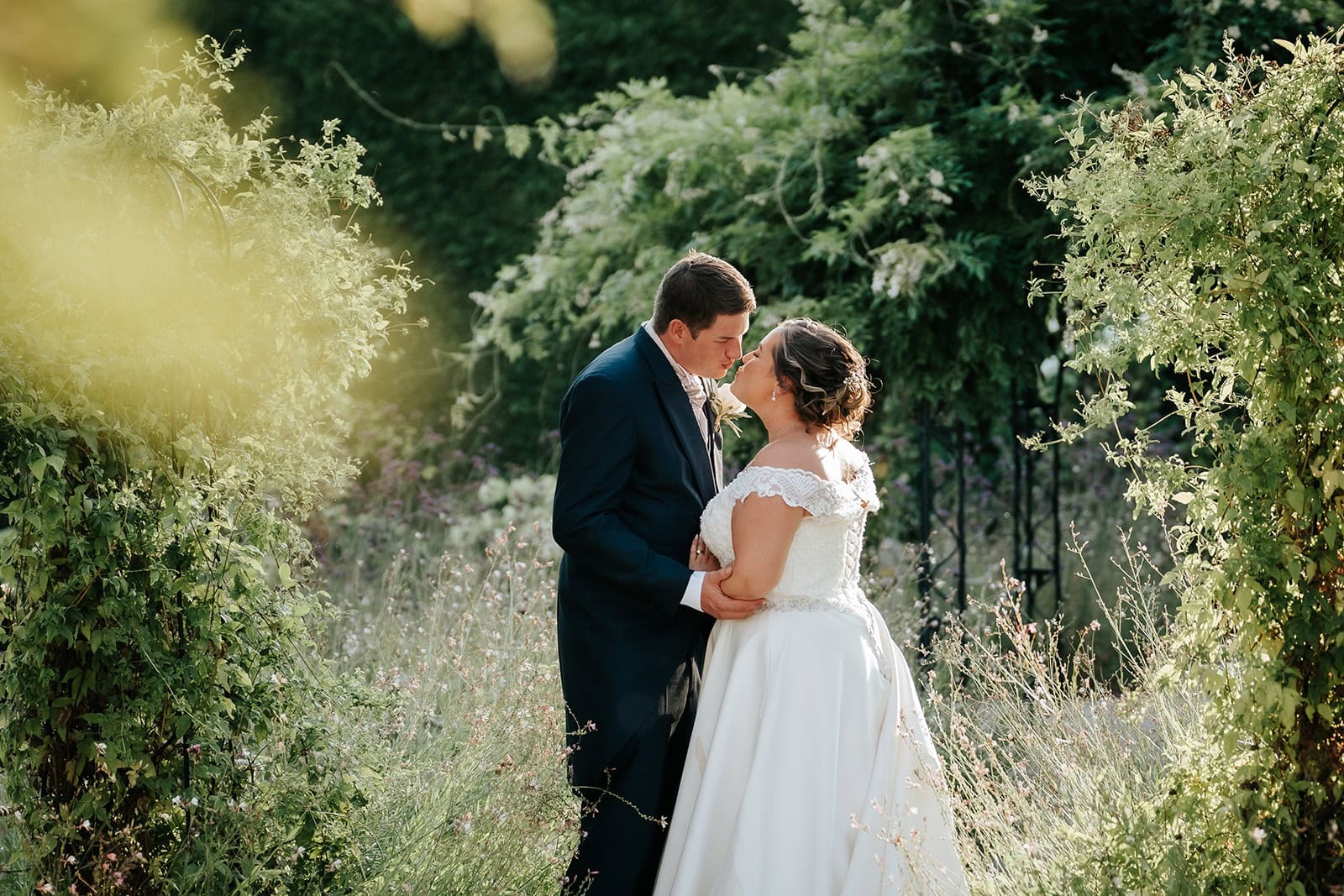 A bride and groom share a quiet moment together in the Victorian Walled Garden at Gaynes Park wedding venue in Epping, Essex. The couple are framed by climbing plants and wild herbaceous borders in soft golden light. Photographed by Tel, Lily & White Photography, Gaynes Park wedding photographer.