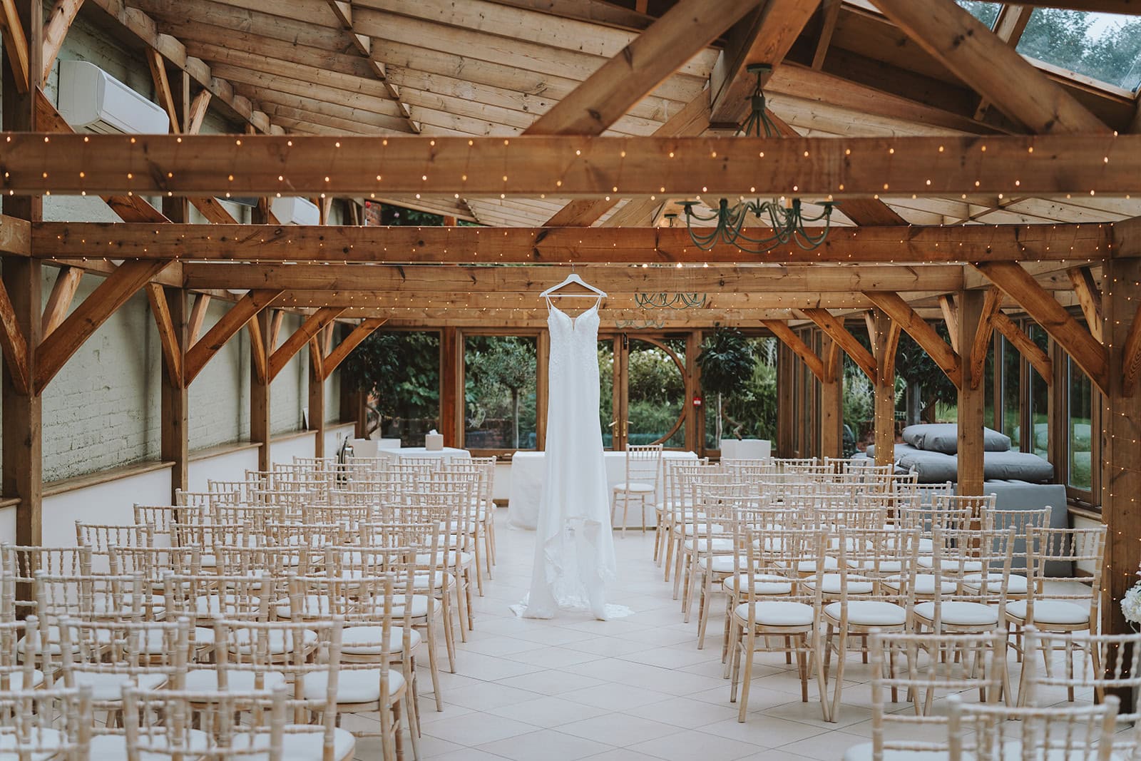 A wedding dress hangs at the end of the ceremony aisle inside the Orangery at Gaynes Park wedding venue in Epping, Essex. Rows of ivory chiavari chairs line the aisle beneath exposed oak beams strung with fairy lights, with the Victorian Walled Garden visible through the floor-to-ceiling windows. Photographed by Tel, Lily & White Photography, Gaynes Park wedding photographer.