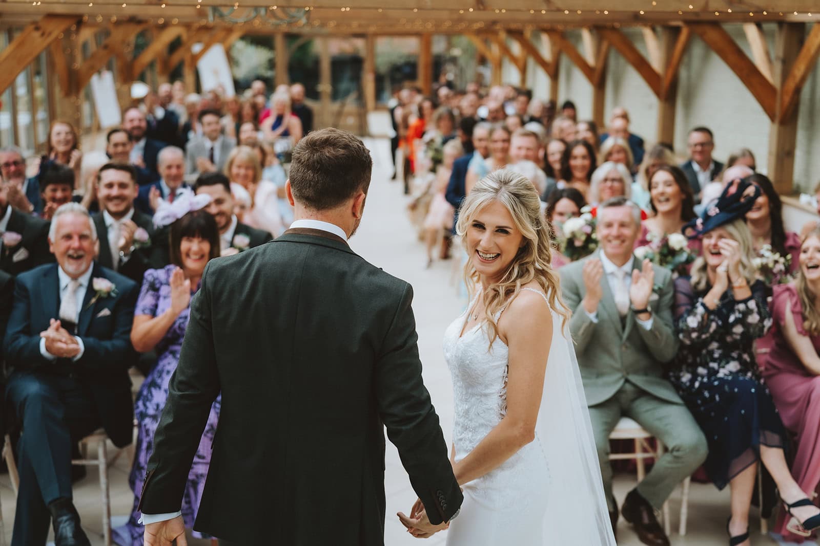 A bride and groom hold hands at the altar inside the Orangery at Gaynes Park, Epping, Essex, as their guests rise in applause and celebration. The full ceremony room of family and friends captured from behind the couple beneath the oak beam and fairy light ceiling. Photographed by Tel, Lily & White Photography, Gaynes Park wedding photographer.