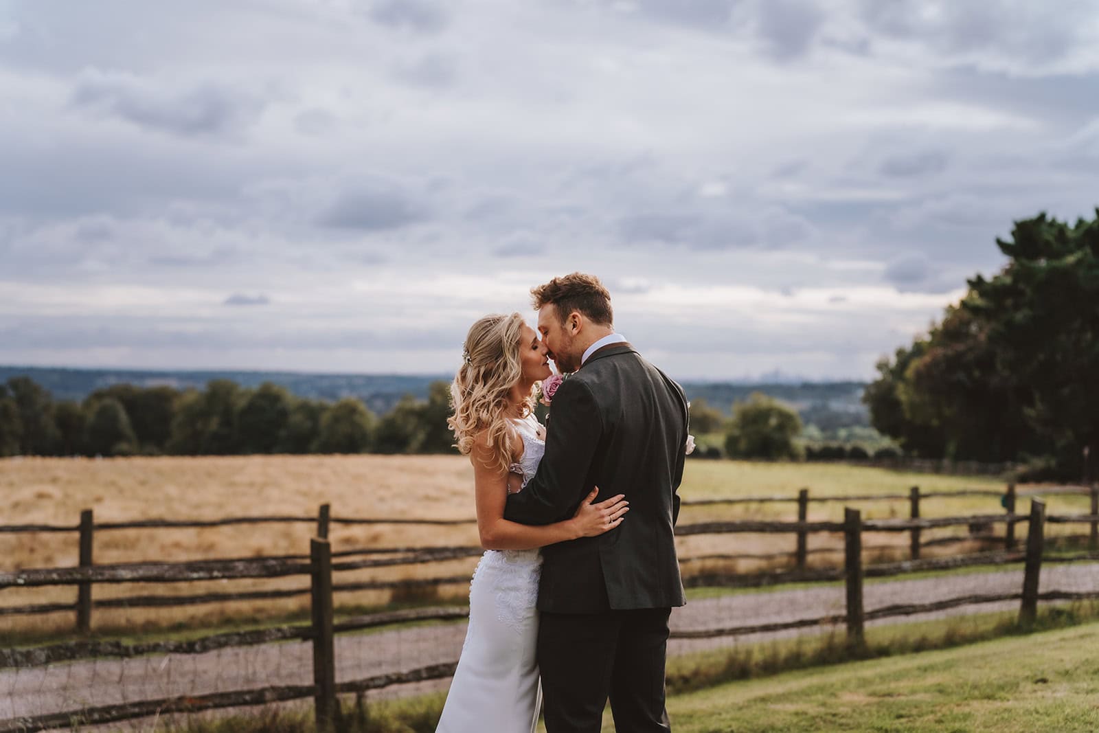 A bride and groom share a kiss at the London Lookout at Gaynes Park wedding venue in Epping, Essex. The couple stand before a sweeping vista of Essex countryside and farmland, with the London skyline faintly visible on the horizon. Photographed by Tel, Lily & White Photography, Gaynes Park wedding photographer.