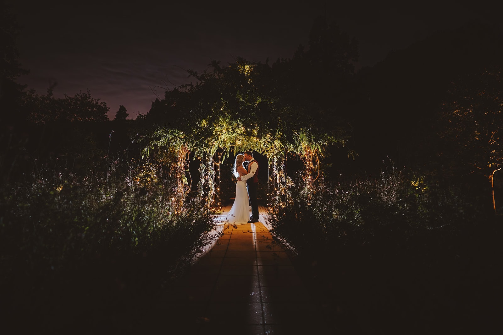 A bride and groom kiss beneath a fairy light-strung pergola arch in the Victorian Walled Garden at Gaynes Park wedding venue in Epping, Essex, photographed at night. The couple are illuminated against the dark sky, surrounded by wild herbaceous borders and twinkling lights threading through the overhanging foliage. Photographed by Tel, Lily & White Photography, Gaynes Park wedding photographer.