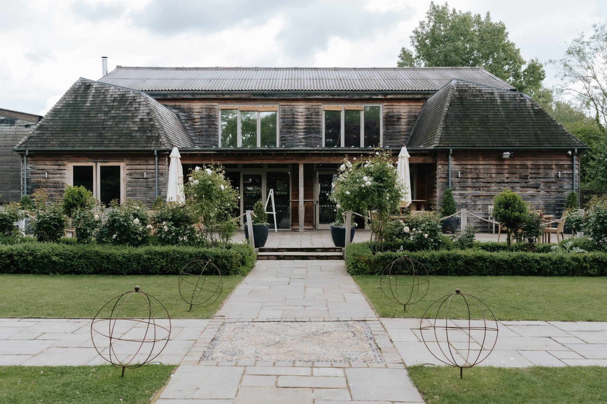 The rustic timber barn exterior at Houchins wedding venue in Coggeshall, Essex, showing the modern glazed upper level and formal garden approach — photographed by Lily & White