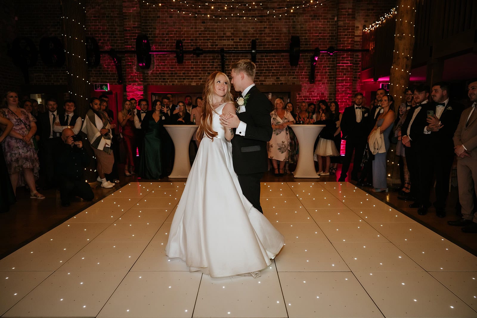 A bride and groom share their first dance on a LED starlit dancefloor in the Mill Barn at Gaynes Park wedding venue in Epping, Essex. The bride looks up at her groom as guests circle the dancefloor, the exposed brick walls lit with pink uplighting and fairy lights above. Photographed by Tel, Lily & White Photography, Gaynes Park wedding photographer.