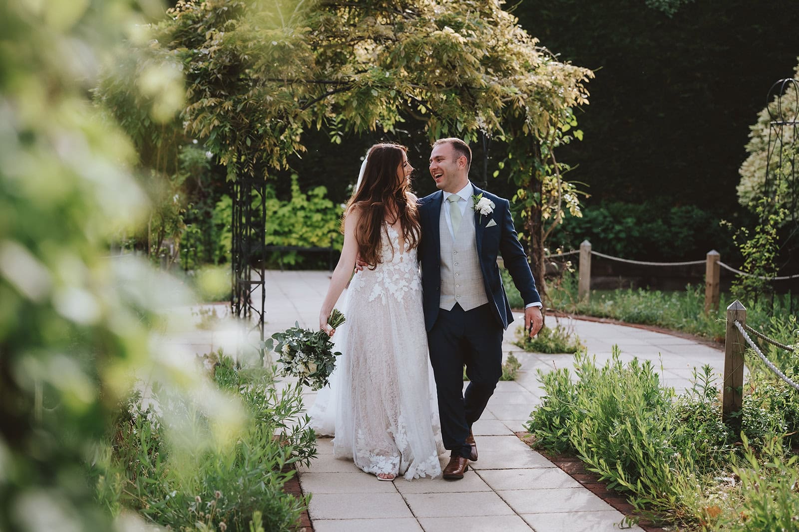 A bride and groom walk together laughing through the Victorian Walled Garden at Gaynes Park, Epping, Essex. Lush green borders frame the stone pathway as the couple share a relaxed, natural moment. Photographed by Tel, Lily & White Photography, Gaynes Park wedding photographer.