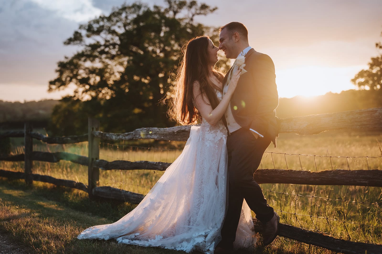 A bride and groom stand close together at golden hour by a rustic wooden fence at Gaynes Park, Epping, Essex. Warm sunset light floods through the trees behind them, casting a cinematic glow across the Essex countryside. Photographed by Tel, Lily & White Photography, Gaynes Park wedding photographer.