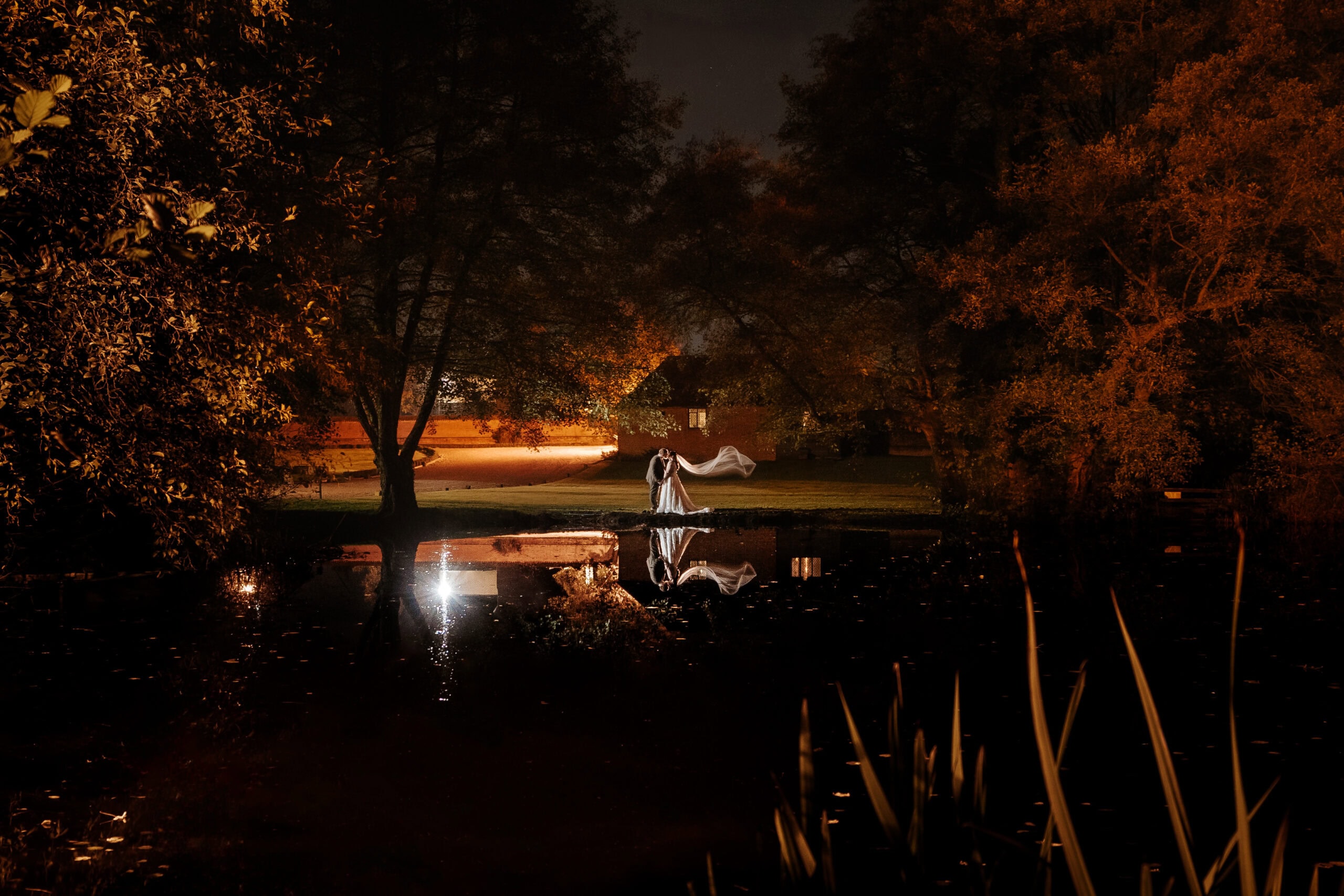 A bride and groom kiss at the edge of the lake at Leez Priory wedding venue in Little Leighs, Essex, photographed at night. The couple are reflected in the still water below, the bride's veil caught in the breeze, with warm amber lighting illuminating the surrounding autumn trees. Photographed by Tel, Lily & White Photography, Leez Priory wedding photographer.