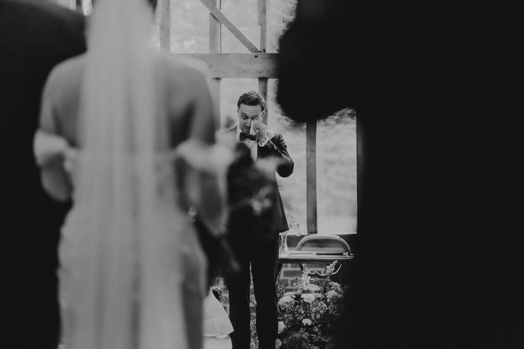 Groom wiping tears during the wedding ceremony, framed by guests in the foreground – natural wedding photography by Tel, Lily & White