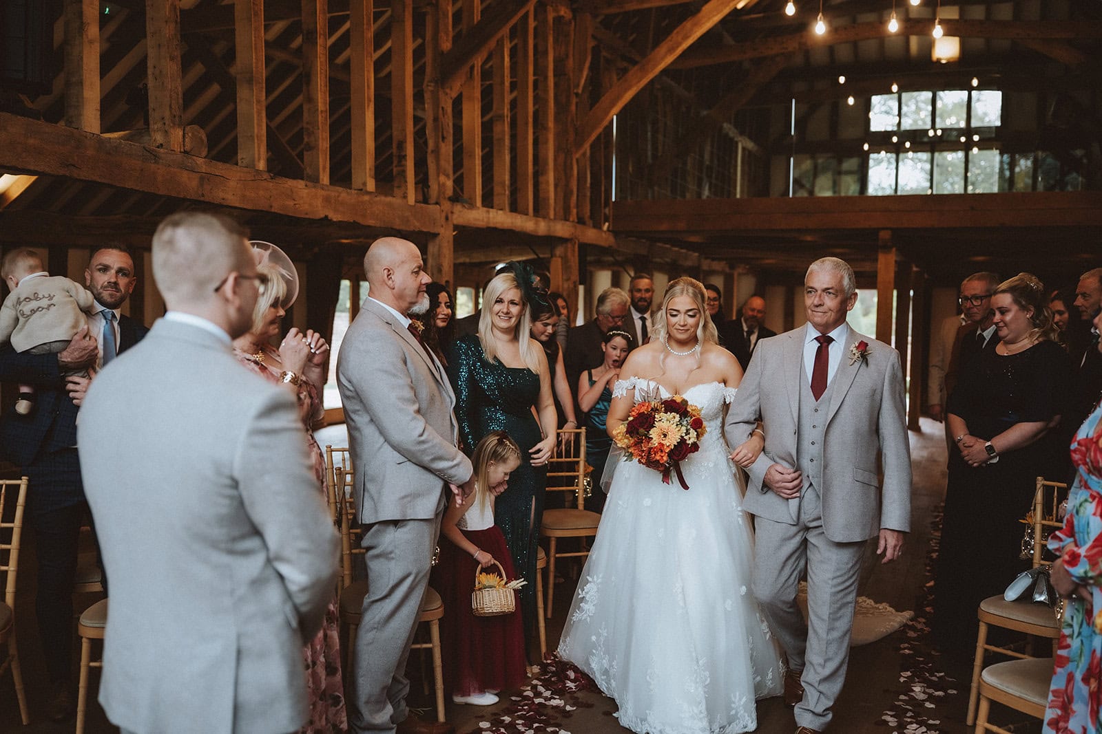 Holly walking down the aisle with her father in the Hay Barn at Blake Hall, Ongar, autumn bouquet of burnt orange and deep red florals, guests lining the aisle, October 2025 – wedding photography by Tel, Lily & White