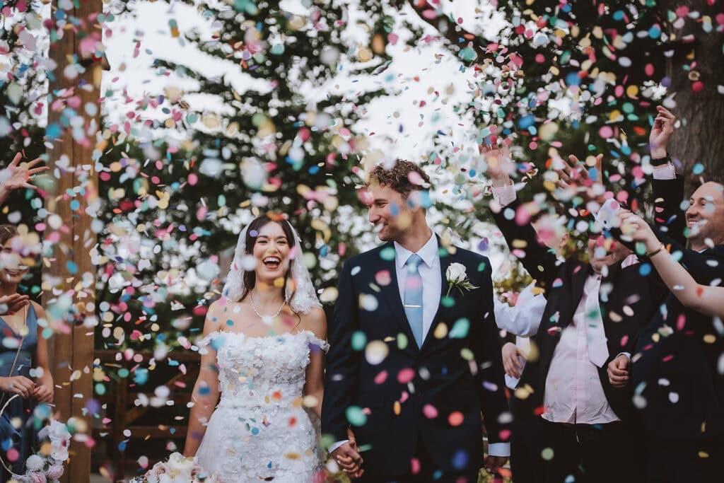 Bride and groom walking through colorful confetti at Friern Manor wedding in Brentwood, Essex — joyful celebration moment captured by Lily & White